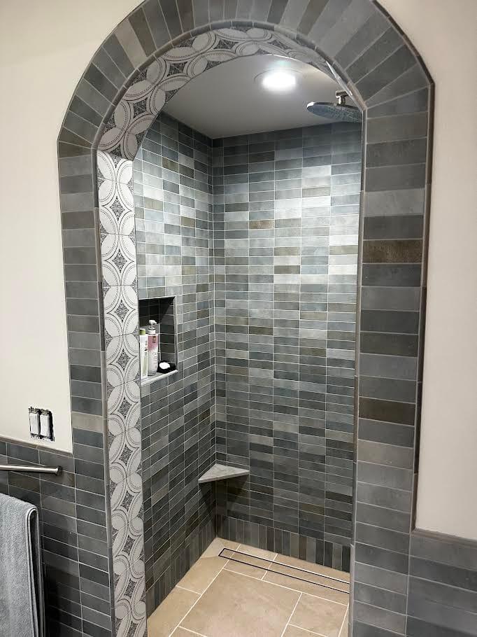 View through a tiled arch into a shower with gray stacked tile, a recessed niche, and a corner seat.